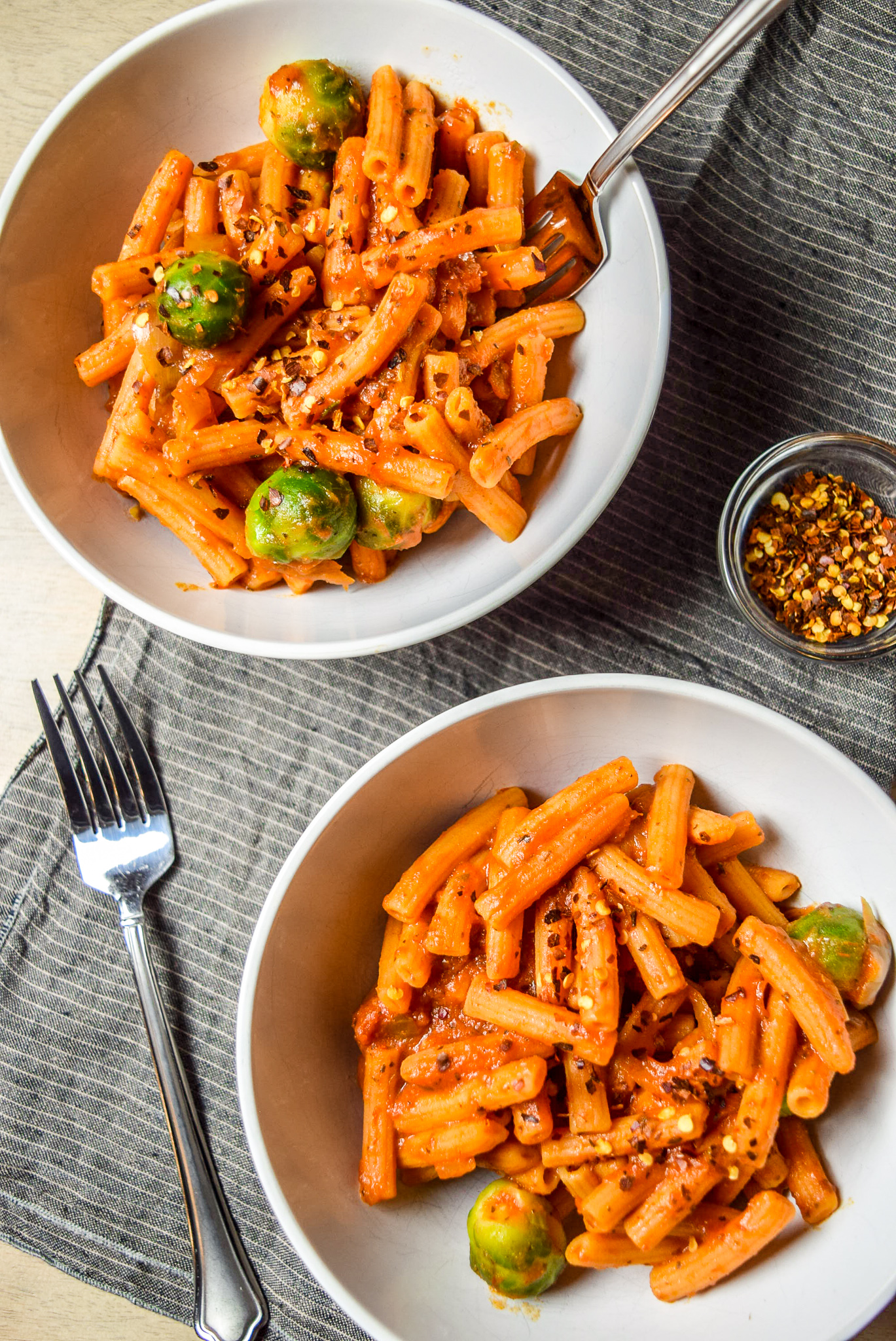 Finished Protein-Packed Red Lentil Pasta with Marinara, Brussel Sprouts, and Caramelized Onions in two bowls with forks, napkin, and chili pepper flakes from top