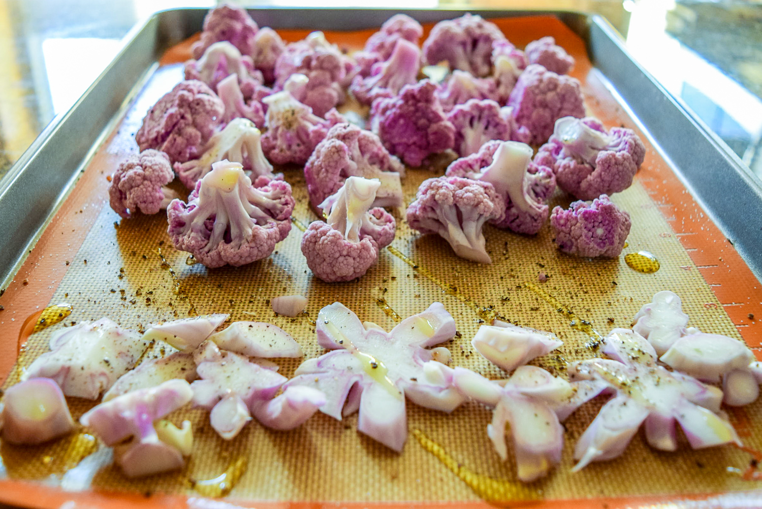 Raw purple cauliflower cut up stem pieces and florets on silicone baking sheet on Calphalon baking sheet from the side with olive oil and salt and pepper