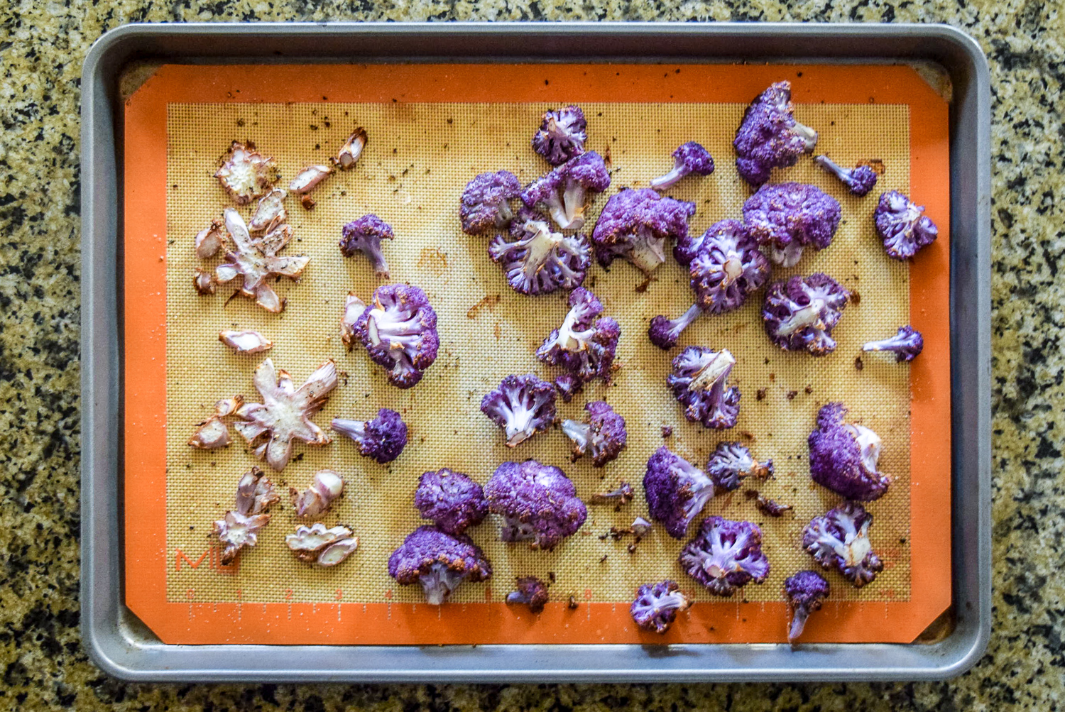 Roasted purple cauliflower florets and stem pieces on silicone baking sheet on calphalon baking sheet from top